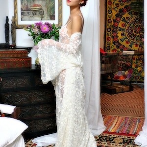 Puede incluir: Un vestido de encaje blanco con mangas largas y una falda fluida. El vestido se usa en una habitaci&oacute;n con una pintura floral, una c&oacute;moda de madera oscura y una alfombra estampada. La est&eacute;tica general es rom&aacute;ntica y bohemia.