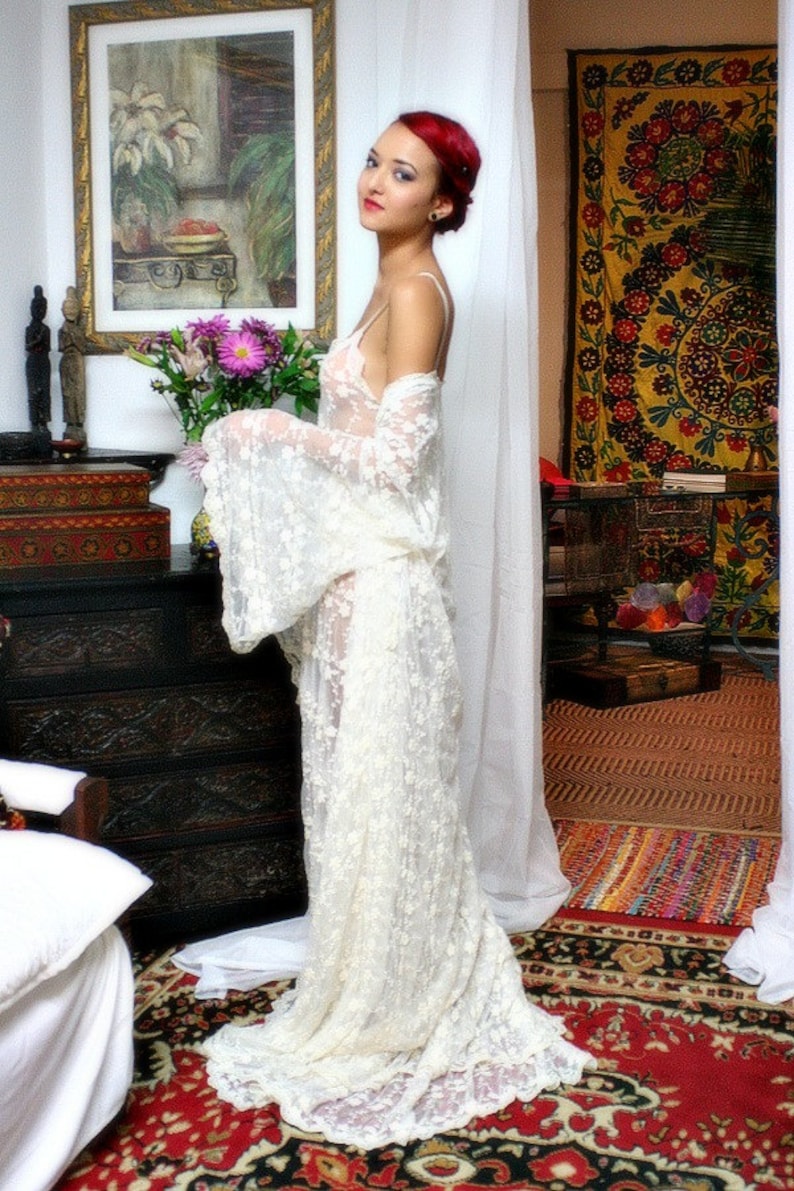 Puede incluir: Un vestido de encaje blanco con mangas largas y una falda fluida. El vestido se usa en una habitaci&oacute;n con una pintura floral, una c&oacute;moda de madera oscura y una alfombra estampada. La est&eacute;tica general es rom&aacute;ntica y bohemia.