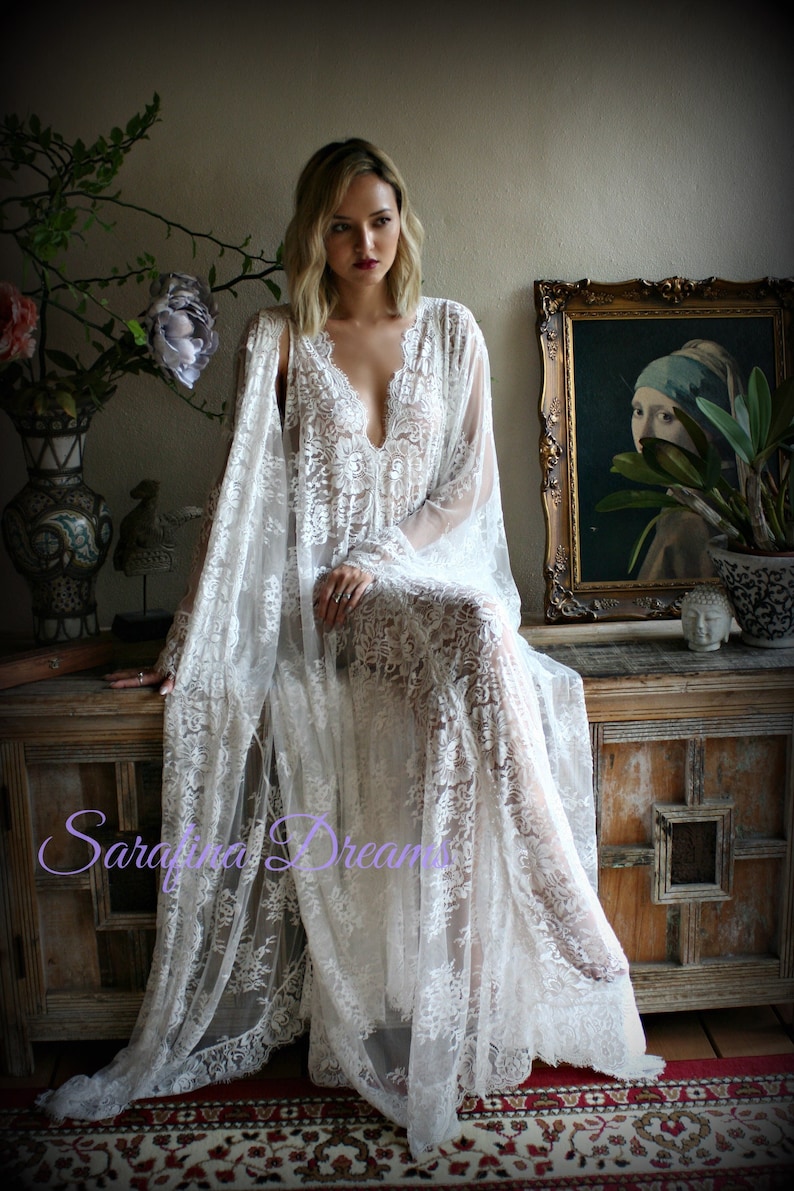 K&ouml;nnte beinhalten: Ein wei&szlig;es Spitzen-Maxikleid mit langen &Auml;rmeln und einem tiefen V-Ausschnitt. Das Kleid ist durchsichtig und hat ein zartes Blumenmuster. Das Model sitzt auf einer Holzbank mit einem Gem&auml;lde im Hintergrund. Der Text "Sarafina Dreams" ist in der linken unteren Ecke des Bildes sichtbar.