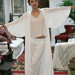 May include: A three-piece white lounge set consisting of a long, open-front duster robe with wide, flowing sleeves, a sleeveless V-neck top, and matching wide-leg trousers with an elasticized waistband, all crafted from a lightweight, solid white fabric.