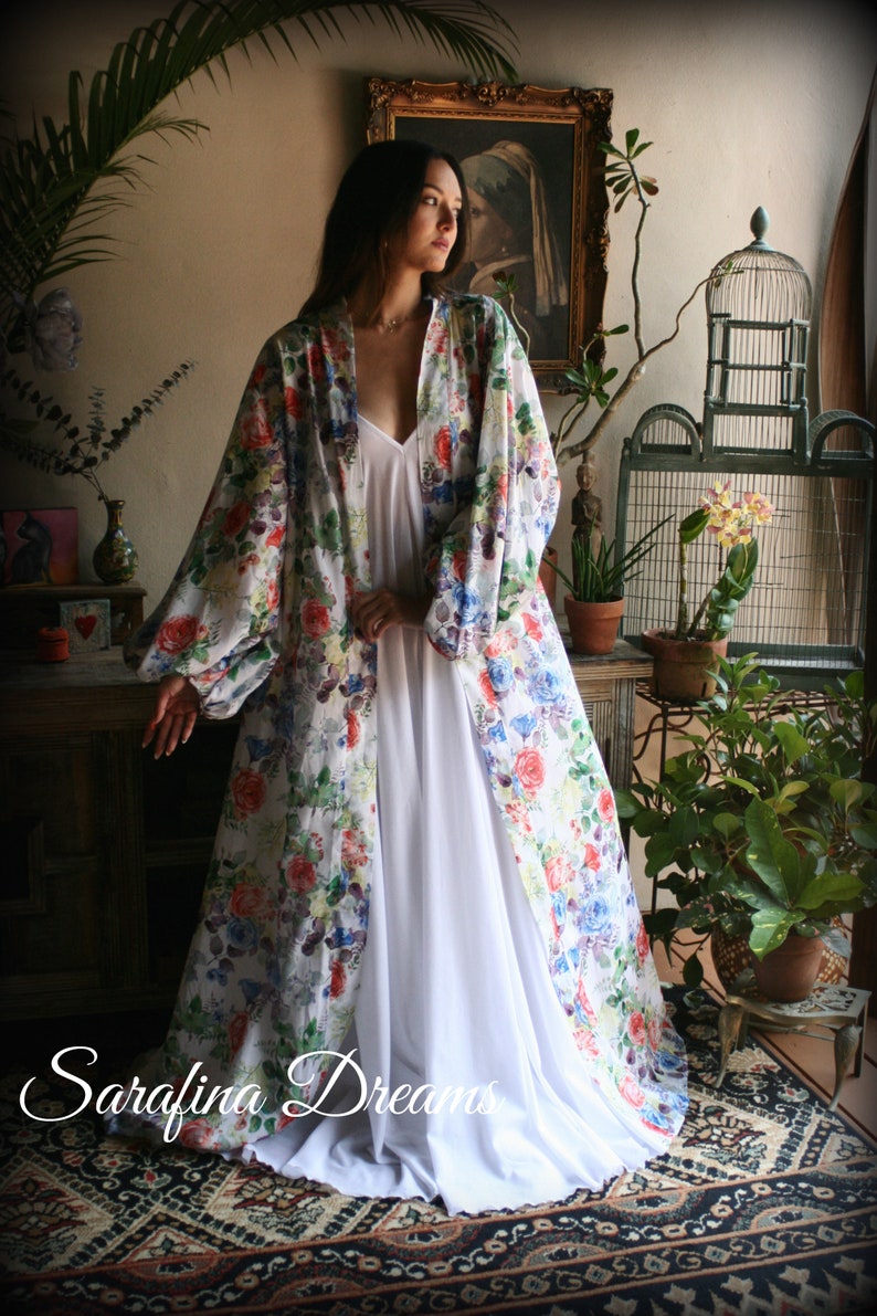 May include: A white floral print robe with long sleeves and a white slip dress underneath. The robe has a loose fit and a V-neckline. The model is standing in a room with plants and a birdcage. The text "Saralina Dreams" is visible in the bottom left corner of the image.