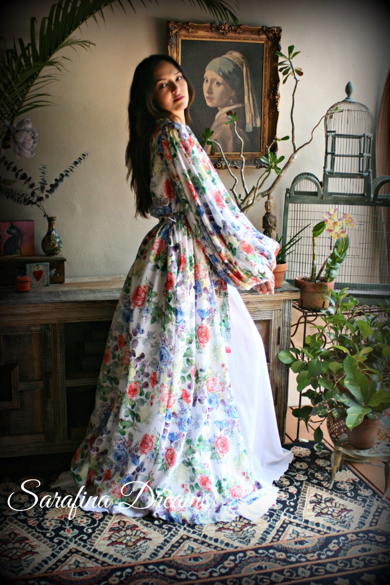 May include: A woman wearing a long, white floral print robe with puffed sleeves. The robe has a white sash tied at the waist. The woman is standing in a room with a painting on the wall and a birdcage in the background.