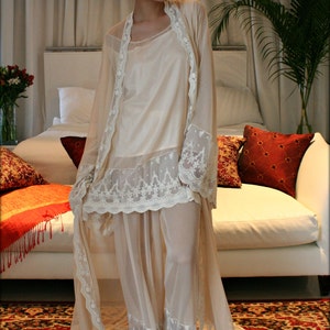 May include: A white lace robe with a sheer overlay and a delicate floral pattern. The robe is worn over a white slip dress. The model is standing on a patterned rug in front of a white couch.