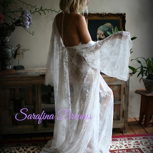 K&ouml;nnte beinhalten: Ein wei&szlig;es Spitzenkleid mit langen &Auml;rmeln und flie&szlig;ender Silhouette. Das Kleid ist durchsichtig und mit zarten Blumenstickereien verziert. Das Model steht vor einem Spiegel, und der Text "Sarafina Dreams" ist in der linken unteren Ecke des Bildes zu sehen.