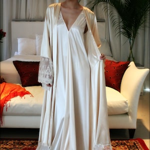 May include: A white satin robe with lace trim and a matching slip. The robe has a V-neckline and long sleeves. The model is wearing the robe and slip and has red hair.