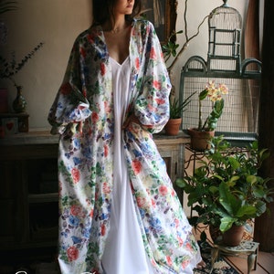 May include: A white floral print kimono with long sleeves and a white slip dress underneath. The kimono has a loose fit and a V-neckline. The model is standing in a room with plants and a birdcage.