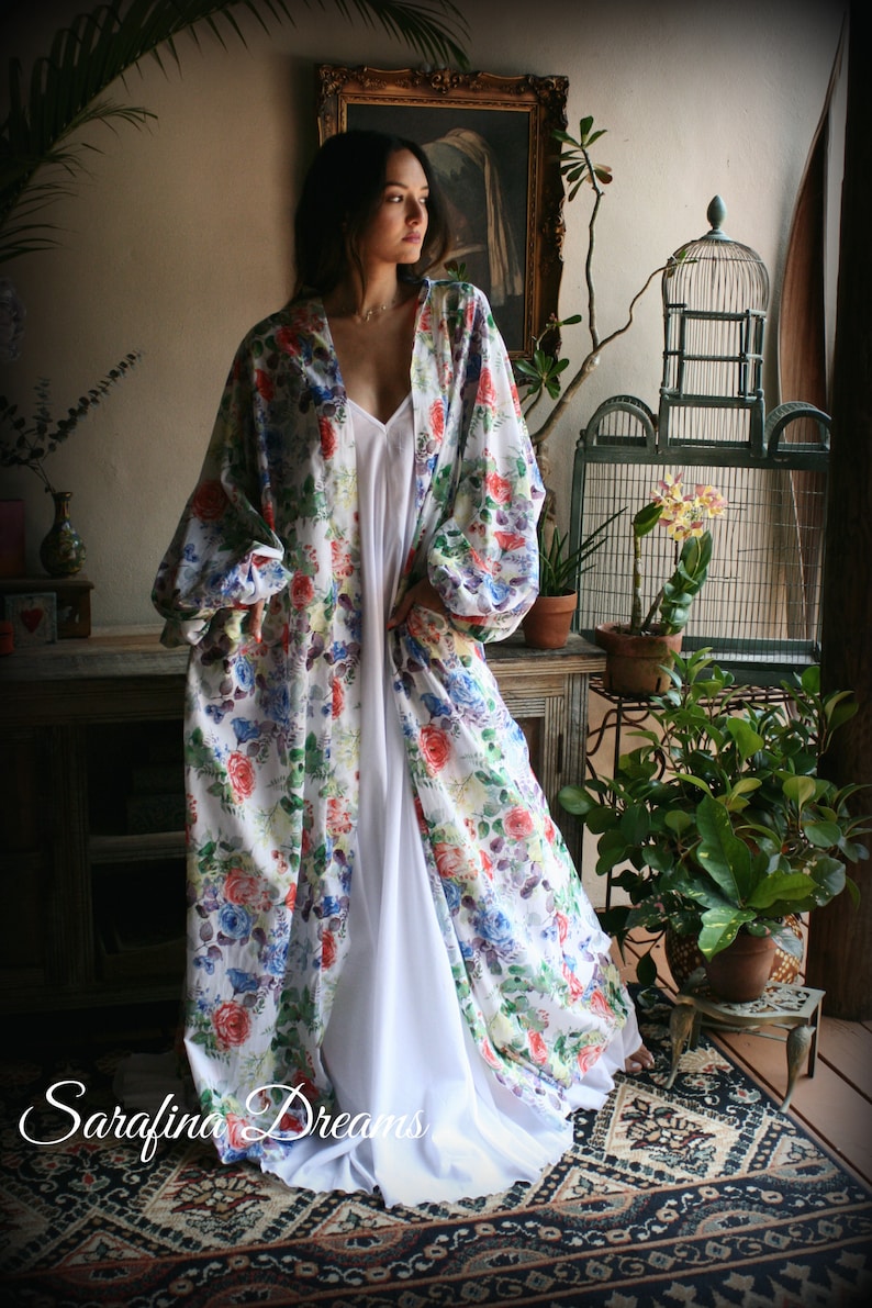 May include: A white floral print kimono with long sleeves and a white slip dress underneath. The kimono has a loose fit and a V-neckline. The model is standing in a room with plants and a birdcage.