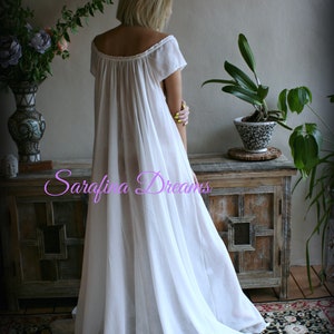 May include: A white cotton nightgown with a scoop neck and short sleeves. The gown is flowing and has a gathered skirt. The model is standing in front of a wooden cabinet with a floral rug on the floor.