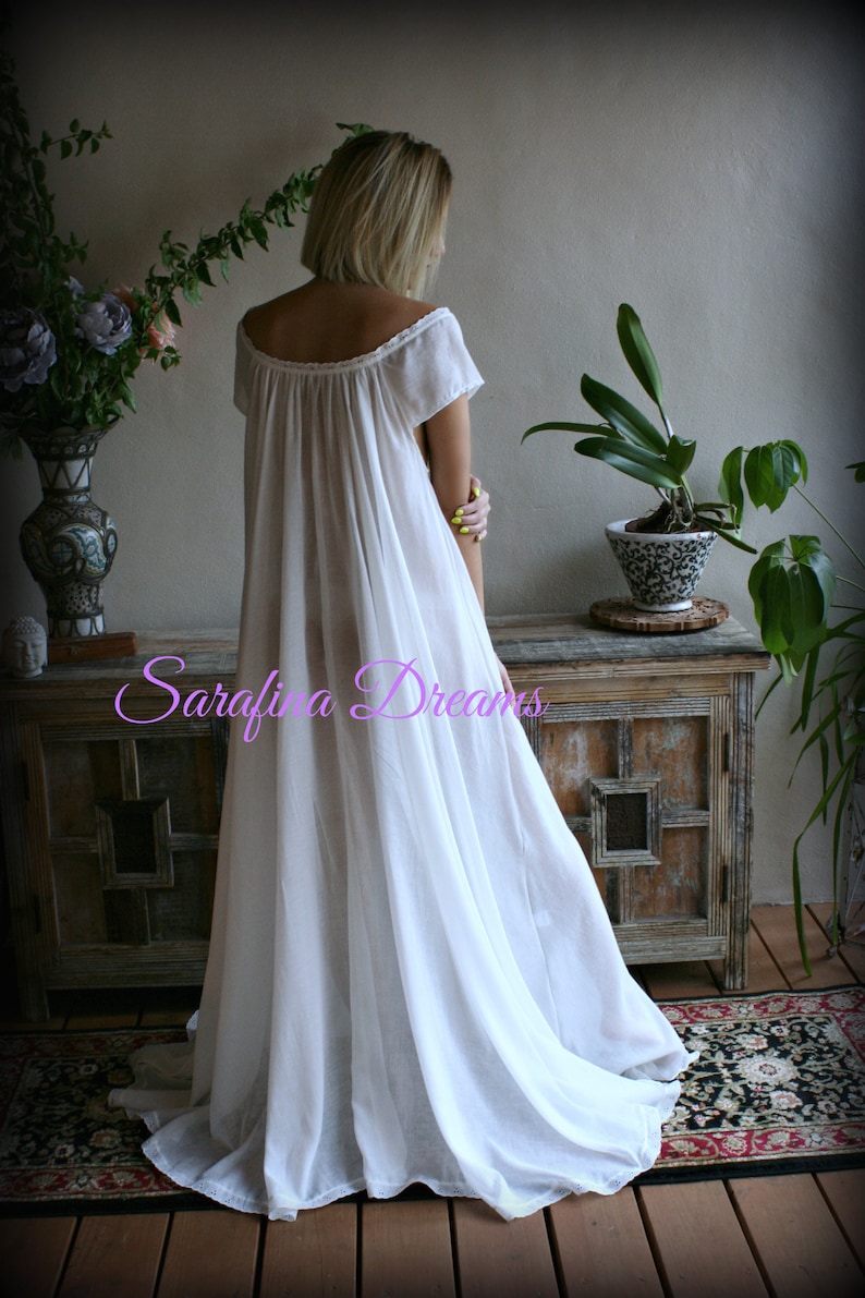 May include: A white cotton nightgown with a scoop neck and short sleeves. The gown is flowing and has a gathered skirt. The model is standing in front of a wooden cabinet with a floral rug on the floor.