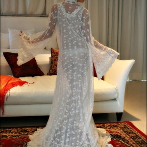 May include: A white lace robe with a long train and bell sleeves. The robe is sheer and has a delicate floral pattern. The model is wearing the robe and standing on a rug.