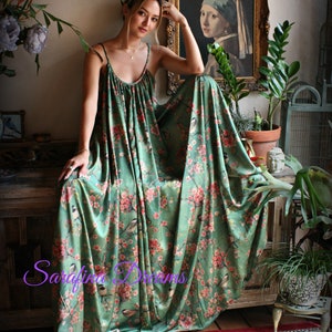 Puede incluir: Vestido largo verde fluido con estampado floral y de p&aacute;jaros. El vestido presenta tirantes finos y un corpi&ntilde;o fruncido. La tela cae sobre una superficie de madera. El vestido est&aacute; adornado con flores rosas y p&aacute;jaros.