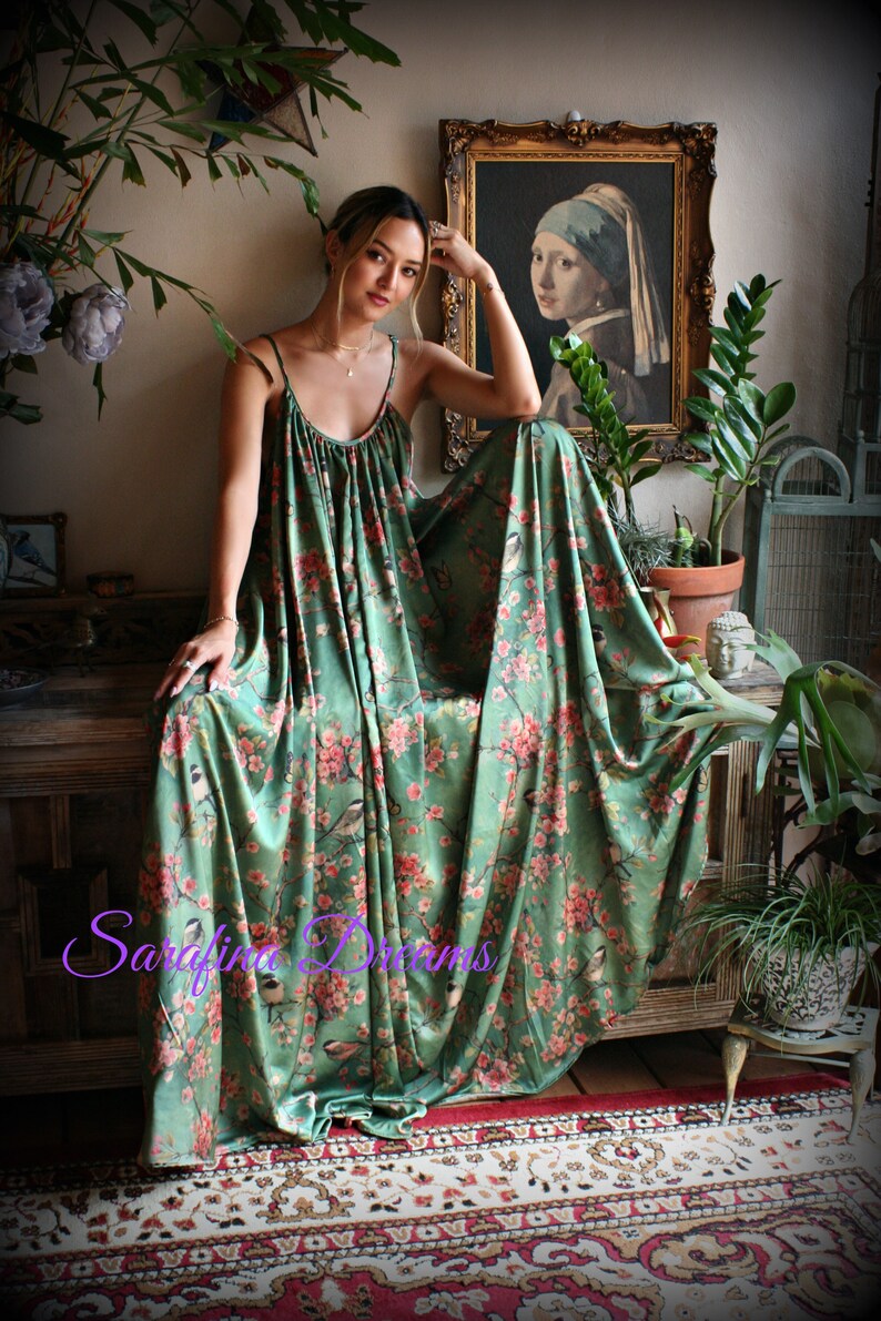 Puede incluir: Vestido largo verde fluido con estampado floral y de p&aacute;jaros. El vestido presenta tirantes finos y un corpi&ntilde;o fruncido. La tela cae sobre una superficie de madera. El vestido est&aacute; adornado con flores rosas y p&aacute;jaros.