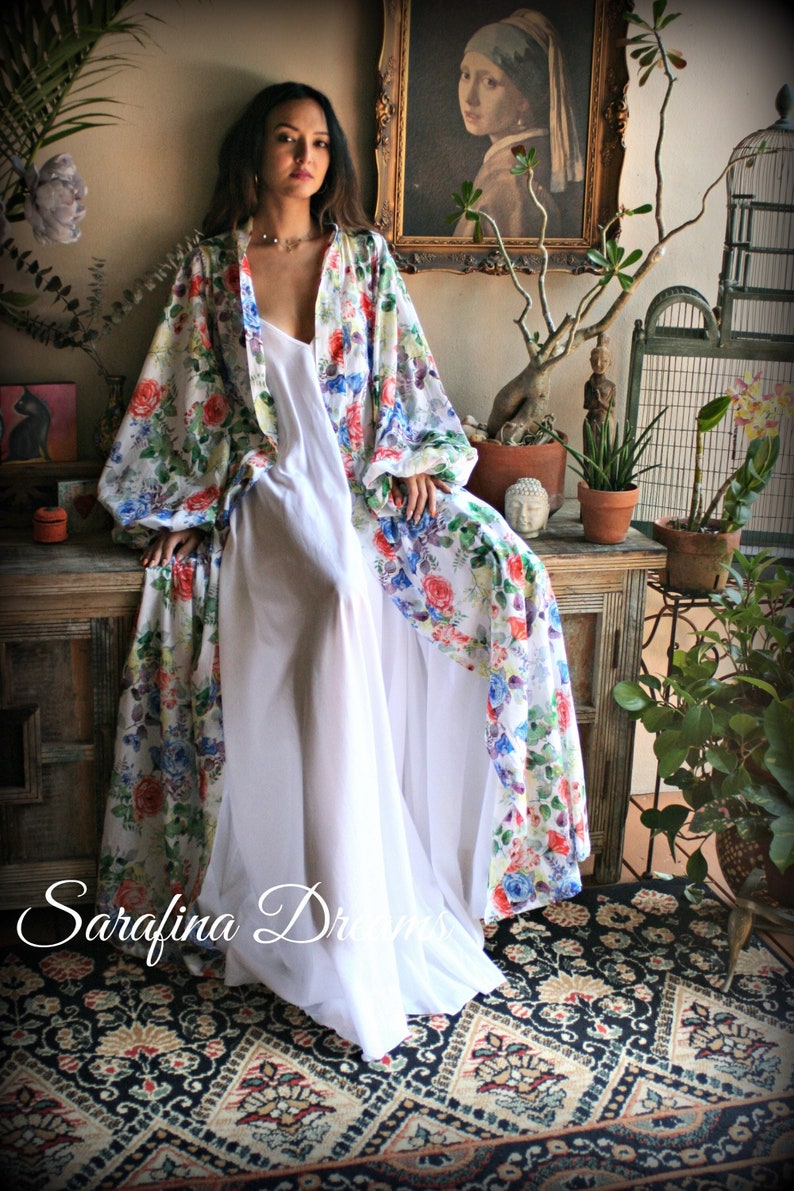 May include: A white maxi dress with a floral print kimono. The kimono has long sleeves and a wide, flowing silhouette. The dress is worn by a person sitting on a wooden chest. The background features a painting, plants, and a birdcage.