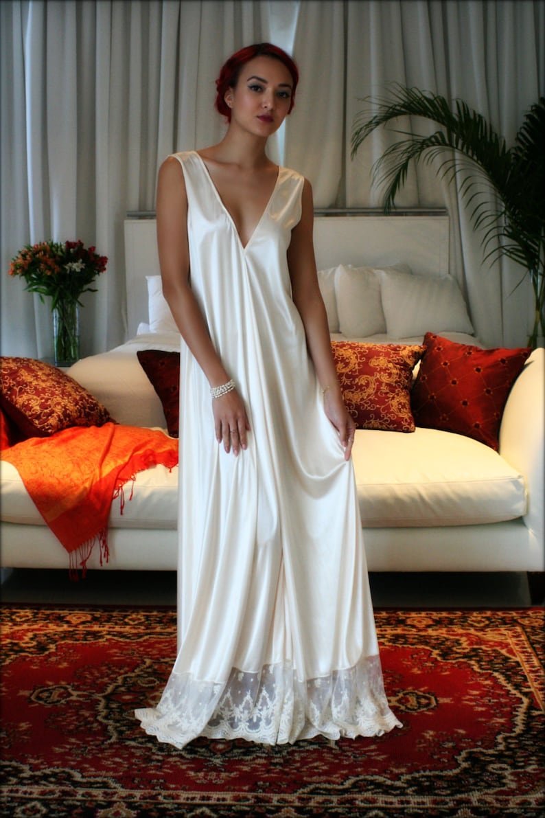 May include: A woman with red hair is wearing a white satin slip dress with lace trim. The dress has a V-neckline and is floor-length. She is standing in front of a white couch with pillows.