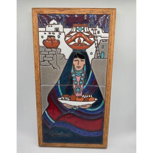 May include: A framed ceramic tile artwork depicting a Native American woman carrying a pot on her head and holding a plate of food. The artwork features a village scene in the background, with a color palette of blue, red, and brown.