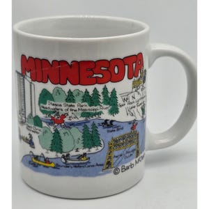 May include: A white ceramic mug featuring a colorful cartoon illustration of Minnesota landmarks and nature. The word MINNESOTA is printed in large red letters at the top. Visible labels include Itasca State Park, Mayo Clinic, and Aerial Lift Bridge.