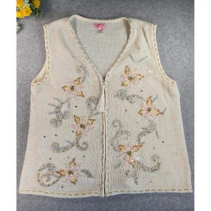 May include: Cream sleeveless knitted vest with a V-neck and a zip. It features floral embroidery in pink, gold, and silver, with sequin details. The edges are trimmed with a contrasting stitch.