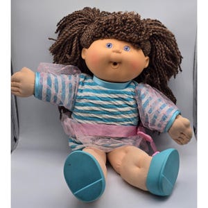 May include: A vintage Cabbage Patch Kids doll with brown yarn hair styled in pigtails. The doll wears a blue and white striped dress with pink accents, blue shoes, and has blue eyes and rosy cheeks. The doll is in a sitting position.
