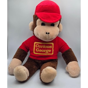 May include: A plush monkey toy with brown fur and a tan face, wearing a red baseball cap and a red shirt. The shirt features a yellow rectangular patch with the text Curious George embroidered in red lettering on the front chest area.