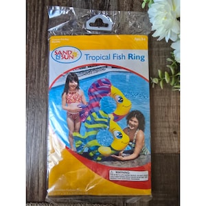Puede incluir: Un flotador inflable para piscina empaquetado, el "Tropical Fish Ring", presenta un diseño de pez colorido con detalles en azul, amarillo y morado. El embalaje incluye el logotipo "SAND N SUN" y una etiqueta de advertencia. El producto está diseñado para edades de 3 a 6 años.