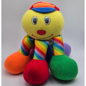 Può includere: Un peluche con una testa rotonda gialla, occhi ricamati e un naso rosso, che indossa un berretto multicolore. Il corpo ha tre gambe con sezioni di tessuto a righe arcobaleno e piedi di peluche arancioni, rossi e verdi.