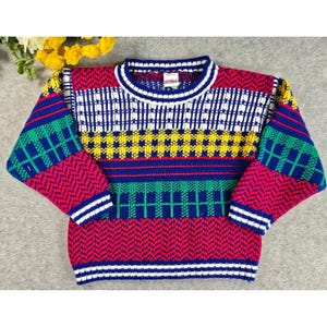 Peut inclure: Un pull en tricot à manches longues présentant un motif géométrique multicolore vif en rouge, bleu, vert, jaune et blanc. Le design comprend des bandes horizontales de motifs chevrons, carreaux et grille avec un col rond et des poignets côtelés.