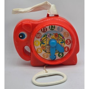 May include: A bright red plastic elephant-shaped clock with a white ribbon handle at the top and a white pull-handle at the bottom. The clock face features numbers one through twelve in various colors and a central illustration of a rabbit on a swing.