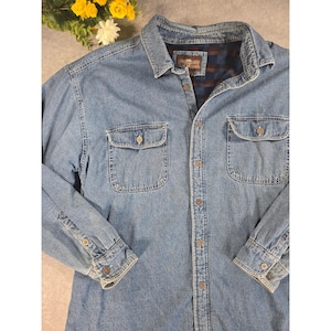 Puede incluir: Camisa vaquera azul claro con dos bolsillos en el pecho y cierre de botones. La camisa tiene mangas largas con puños abotonados y cuello. El forro interior presenta un estampado de cuadros.