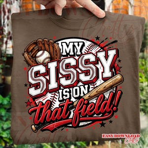 Op de afbeelding: Bruin sweatshirt met een honkbalthema-afbeelding. Het ontwerp toont een honkbal, handschoen en knuppel, met de tekst "MY SISSY IS ON THAT FIELD!" in rood en wit. Het ontwerp bevat sterren en een honkbal.