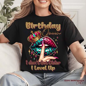 May include: Black t-shirt with a gold glitter design that reads "Birthday Queen." The graphic features a crown, lips with a leopard print design in pink and teal, and a finger to the lips. The text "I don't Get Older I Level Up" is below the lips.