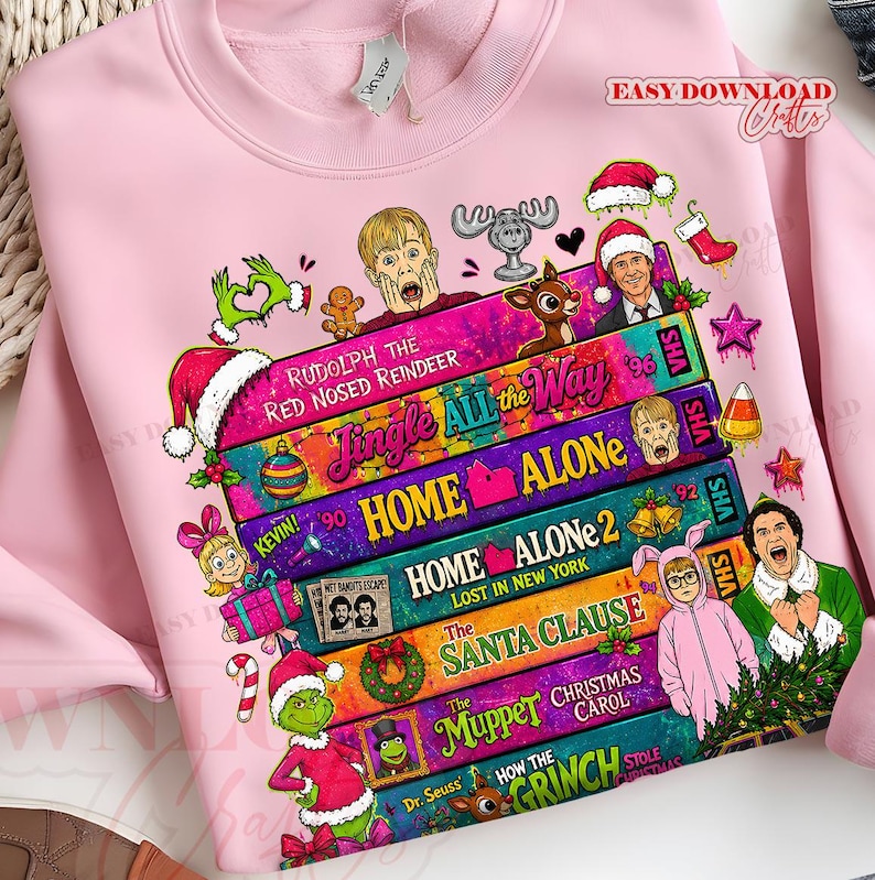 Puede incluir: Una sudadera rosa claro con un estampado gr&aacute;fico de cintas VHS apiladas con t&iacute;tulos de pel&iacute;culas navide&ntilde;as como Home Alone, The Santa Clause y The Muppet Christmas Carol, rodeadas de personajes festivos e iconos navide&ntilde;os.