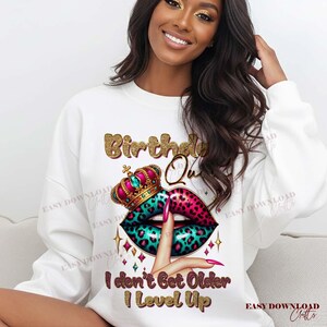 May include: White sweatshirt with a graphic design featuring the text "Birthday Queen" in gold glitter lettering. The design includes a crown, lips with a teal and pink leopard print, and the text "I don't Get Older I Level Up."