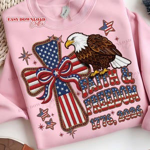 May include: Pale pink sweatshirt with a patriotic design. It features an American flag cross, a bald eagle, and the text "Faith & Freedom 1776, 2026." The design uses red, white, and blue, with star and stripe details.