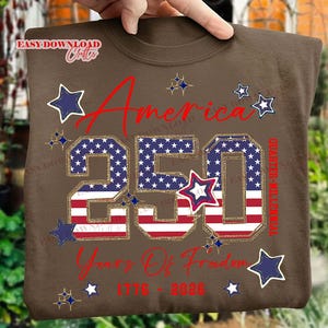 May include: A brown t-shirt with the text "America 250 Years Of Freedom 1776-2026" in red and gold. The number "250" is filled with a stars and stripes pattern, and there are star graphics.