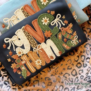 May include: A black decorative pillow with the word "Autumn" in a knitted style, featuring fall-themed elements like leaves, acorns, and flowers. The pillow has a bow and a leopard print accent. The background includes a light blue pillow and a leopard print fabric.