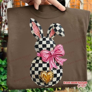 May include: Brown sweatshirt featuring a whimsical bunny design. The bunny has a black and white checkered pattern with pink inner ears, a pink bow, and a gold heart. The design is centered on the front of the sweatshirt.