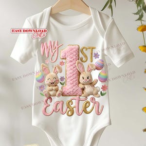 May include: White baby bodysuit with a pink and gold "My 1st Easter" design. The design includes a large pink number 1, two cartoon bunnies, Easter eggs, and floral accents. The bodysuit is hanging on a wooden hanger.