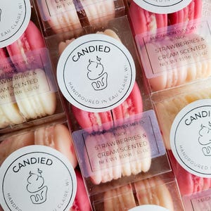 May include: Multiple clear plastic packages containing wax melts shaped like pink and cream-colored macarons. Each package features a round white label with the text Candied Handpoured in Eau Claire, WI and a rectangular label stating Macaroon Wax Melts 6 Pack Strawberries & Cream Scented Net Wt 3.5 oz.