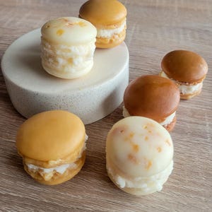 May include: Six macarons with different colored shells and white filling. The macarons are arranged on a white surface and a wooden surface.