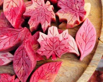 May include: A collection of leaf-shaped wax melts in shades of red and pink, arranged on a wooden tray. The melts feature detailed vein patterns, creating a realistic autumn theme. The tray has a natural wood grain.