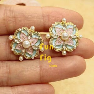 Könnte beinhalten: Zwei blütenförmige Ohrringe mit goldfarbenem Rand, mit hellblauem und rosa Emaille-Design. Jeder Ohrring hat eine zentrale Perle und ist mit klaren Kristallen und kleineren Perlen verziert. Der Text "fun fig Crafts" ist sichtbar.