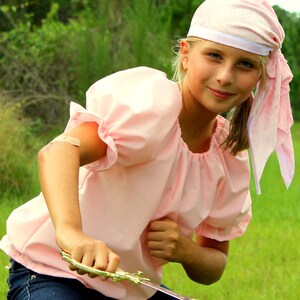 Può includere: Una giovane persona indossa una blusa rosa e un bandana rosa e bianco, tenendo una spada giocattolo dorata. La persona è in piedi in un campo erboso.