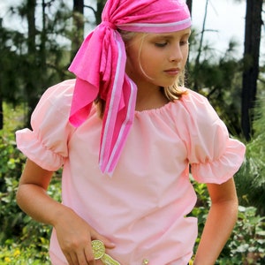 Può includere: Una giovane persona indossa una camicia da pirata rosa e un bandana rosa con bordi bianchi. Sta tenendo una spada giocattolo dorata.