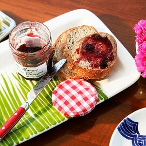 May include: A white plate with a green grass design features a knife, a jar of jam, a lidded jar, and two slices of bread with jam. A small silver dish with a stick of butter is in the background. A small bowl with pink carnations is on the right side of the image.