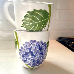 May include: Two white ceramic mugs with floral designs. The top mug has a green leaf design, and the bottom mug has a purple hydrangea design.
