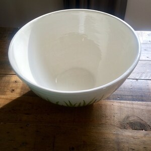 May include: White ceramic mixing bowl with a green leafy design around the bottom edge. The bowl is empty and sitting on a wooden surface.