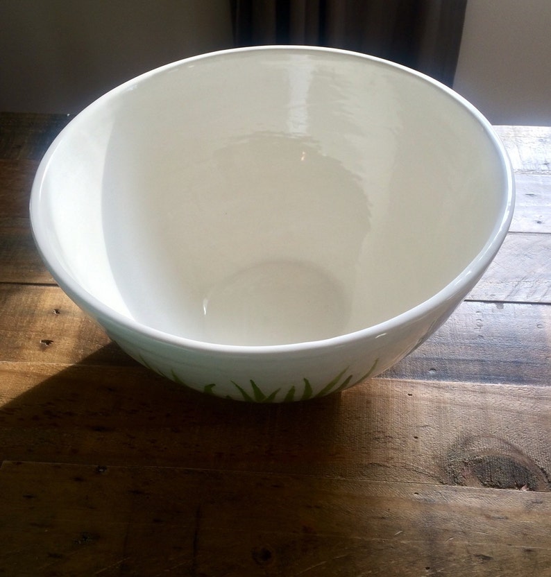 May include: White ceramic mixing bowl with a green leafy design around the bottom edge. The bowl is empty and sitting on a wooden surface.