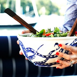 May include: A white ceramic bowl with a blue and white octopus design, filled with a green salad. The bowl is being held by a person with red nail polish.