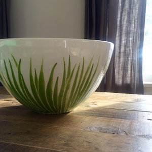 May include: A white ceramic bowl with a green grass design. The bowl is sitting on a wooden table.