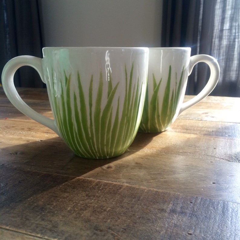 May include: Two white ceramic mugs with green grass designs. The mugs have a rounded shape and a handle on the side.